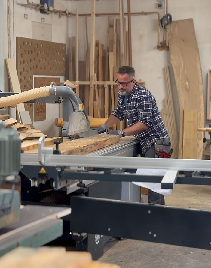 Ein Handwerker in einem karierten Hemd, der eine große Holzsäge bedient und ein Holzbrett in einer Werkstatt mit Werkzeugen und Holz zuschneidet.