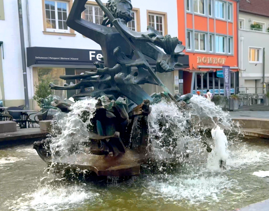 Ein lebhafter Brunnen mit einer skulptierten Figur, umgeben von sprudelndem Wasser, auf einem belebten Marktplatz mit Gebäuden im Hintergrund.