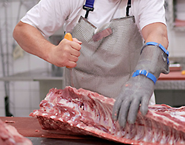 Ein Metzger mit Schutzhandschuhen und Schürze, der ein großes Stück Fleisch auf einem Schneidetisch in einem sauberen Verarbeitungsraum zerteilt.