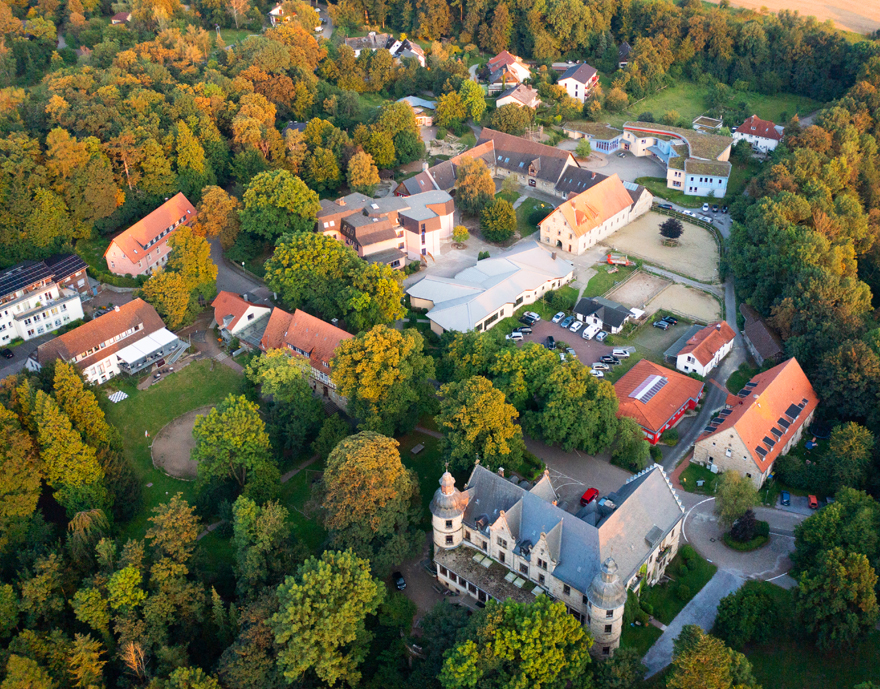 Eine Luftaufnahme von Schloss Hamborn, die eine Ansammlung von Gebäuden zeigt, umgeben von üppigem Grün und einem angrenzenden Feld.